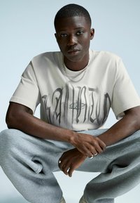 Młody mężczyzna z krótkimi włosami, ubrany w biały t-shirt z grafiką, srebrny naszyjnik i pierścienie, siedzi na podłodze w pozycji skrzyżowanej na tle jasnym, jednolitym.