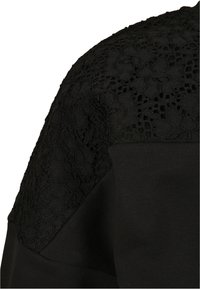 Détail d'épaule en tissu noir combinant tricot uni et motif de dentelle florale avec des ouvertures en résille.