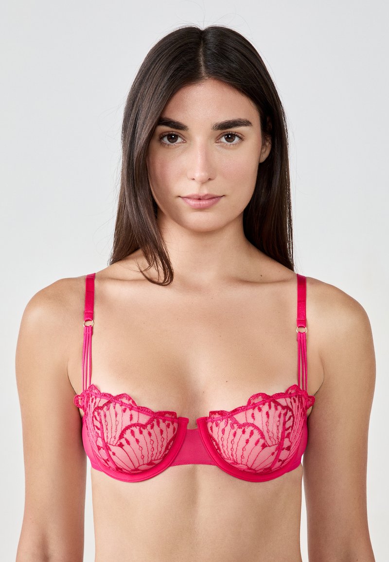 Różowa koronkowa biustonosz z przezroczystymi panelami, falbaniastymi krawędziami i delikatnymi detalami. Posiada regulowane ramiączka i gładki pas.