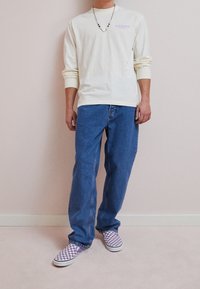 Persoon in een crèmekleurig shirt met lange mouwen, blauwe spijkerbroek, paarse en witte geruite instappers, en een kralenketting met een bloemenhanger.