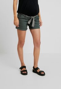 Mörkgröna shorts med uppvikta fållar, prydda med ett svart och vitt randigt bälte. Kombineras med svarta sandaler med justerbara remmar.