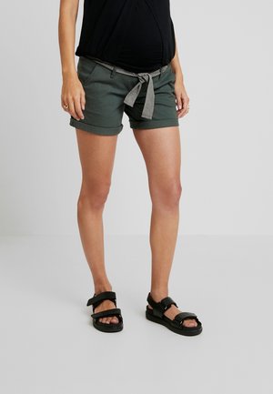 Ciemnozielone szorty z podwiniętymi nogawkami, z czarno-białym paskiem w formie paska do przewiązania. W zestawie z czarnymi sandałami z regulowanymi paskami.