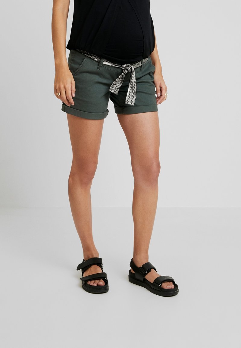 Mörkgröna shorts med uppvikta fållar, prydda med ett svart och vitt randigt bälte. Kombineras med svarta sandaler med justerbara remmar.