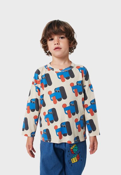 Long-sleeve beige shirt featuring colorful cartoon dog patterns in blue, black, and red, with a rounded neckline. Worn with blue jeans.