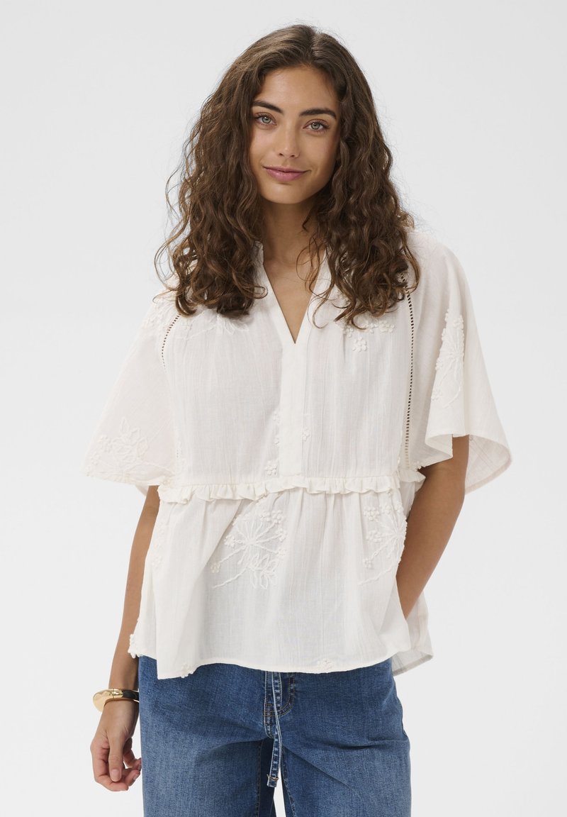 Femme aux cheveux bouclés portant un chemisier blanc brodé ample avec des manches évasées et un jean bleu, debout avec une main dans la poche.