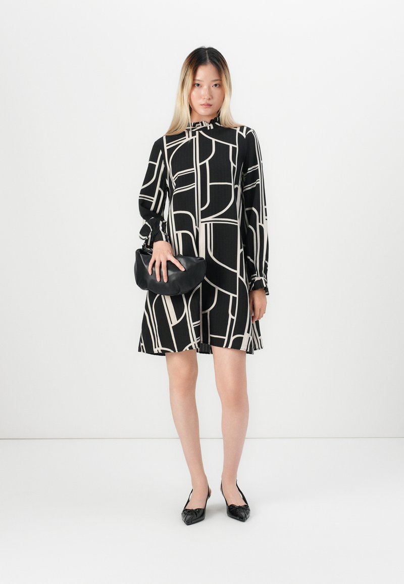 Mujer vestida con un vestido de patrón geométrico en blanco y negro, sosteniendo un clutch negro, de pie contra un fondo blanco plano.