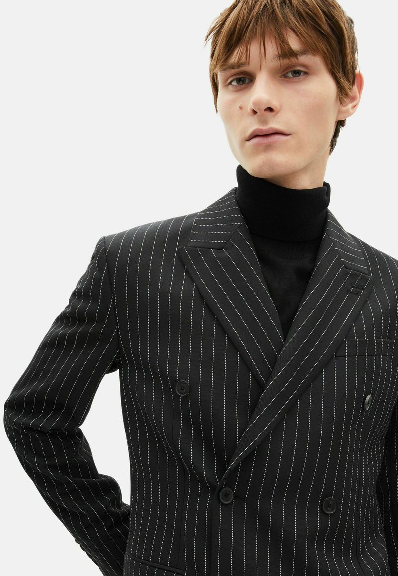 Veste noire à double boutonnage à rayures fines blanches, avec revers en cranté, poches latérales et tissu texturé. Portée sur un col roulé noir.