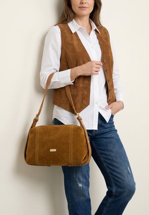 Kvinde iført hvid skjorte, brun ruskindsvest, blå jeans, der holder en matchende brun ruskind skuldertaske med guldfarvet hardware.
