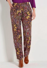 Frau trägt eine Hose mit Paisleymuster in Lila-, Braun- und Grüntönen, kombiniert mit braunen Stiefeletten und einem lila Oberteil.