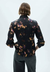 Blouse noire à motifs floraux violets et orange, manches longues, col à boutons. Texture lisse et coupe ajustée.