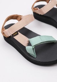Sandalias Teva con una suela texturizada negra, correas ajustables en beige y verde menta, y una suela de goma suave para mayor comodidad.