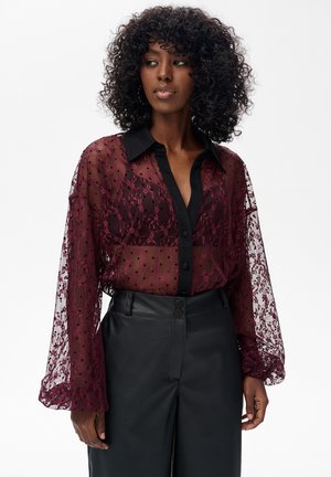 Blouse van kant in bordeauxrood met een zwarte kraag, lange transparante mouwen en knoopsluiting. Gecombineerd met zwarte leren hoge taille broek.