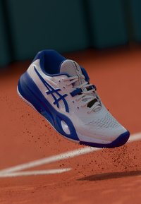 Chaussure de sport blanche et bleue avec une tige en maille, des détails texturés et une semelle en caoutchouc, conçue pour la performance sportive sur un court en terre battue.