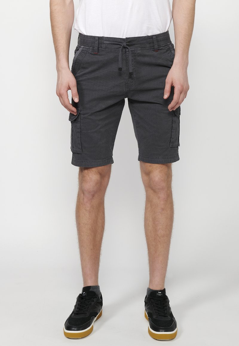 Man som bär mörkgrå cargo shorts, vit t-shirt, svarta sneakers, står mot en vit bakgrund, med händerna vid sidan av benen.