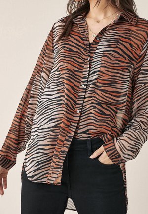 Vrouw draagt een doorschijnende blouse met lange mouwen en tijgerstrepen in oranje, zwart en beige, gecombineerd met een zwarte broek, één hand in de zak, zichtbare gelaagde gouden kettingen.