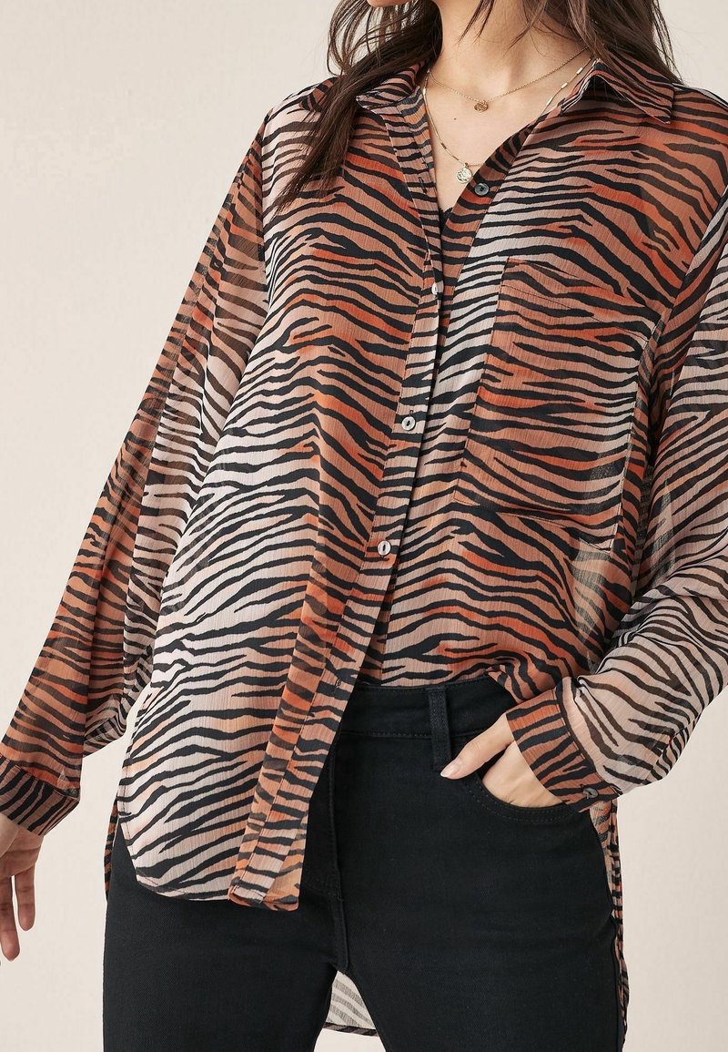 Femme portant un chemisier transparent à manches longues à rayures tigre orange, noir et beige, avec un pantalon noir, une main dans la poche, des colliers en or superposés visibles.