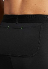 Zwarte sportshorts met een vlakke tailleband, voorzien van groene stikselaccenten en een gladde textuur. Close-up van het achterontwerp.