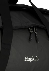 Schwarze Sporttasche mit verstellbaren Trägern, Reißverschluss und schwarzen Kordeln. Besitzt ein weißes "Haglöfs"-Logo auf der Vorderseite.