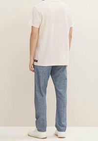 Mężczyzna w gładkiej białej koszulce, niebieskich lekkich spodniach i białych sneakersach, stojący na neutralnym tle tyłem.