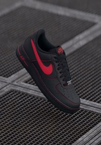 Black leather sneaker with red accents, featuring a perforated toe box, black laces, and a rubber sole. Nike logo and "AIR" detail visible.