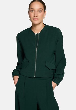 Femme portant une veste zippée vert foncé avec poches à rabat et pantalon assorti, debout les mains dans les poches devant un fond uni.