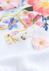 Hvidt stof med farverigt blomstermønster, lyserødt stofblomst og gylden broderet sløjfe samt tekst, der lyder "NEVER".