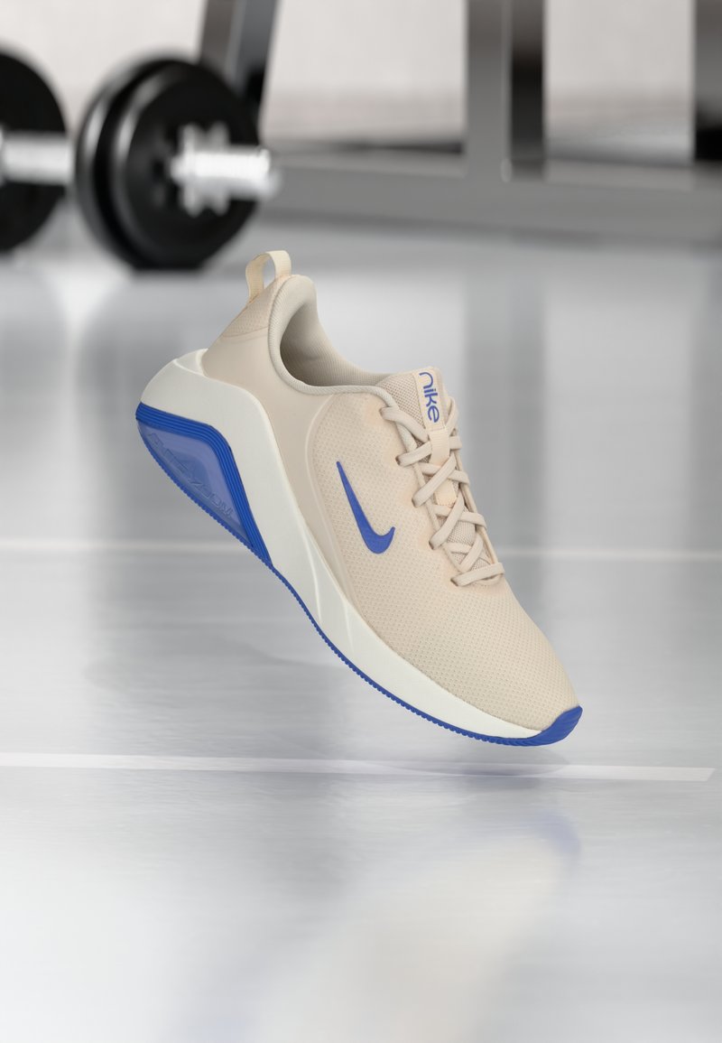 Beige Sportlich-Schuh mit einer strukturierten Oberfläche, blauen Akzenten am Nike-Logo und der Sohle sowie einer gepolsterten weißen Zwischensohle, die für Komfort ausgelegt ist.