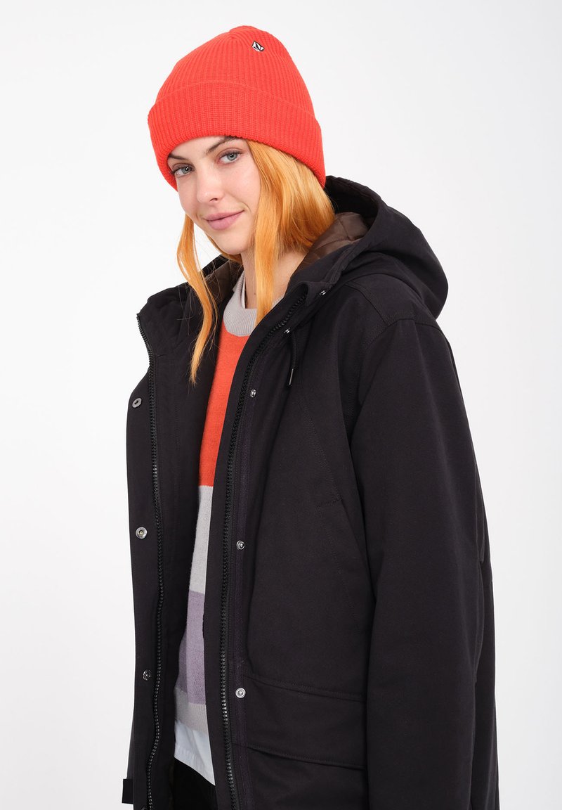 Giacca nera con cappuccio, chiusura a zip e bottoni a pressione, abbinata a un maglione a righe arancione, grigio e bianco, e a un berretto di maglia rosso brillante.