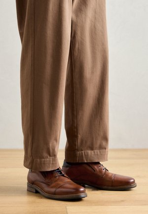 Scarpe in pelle marrone con finitura liscia, punta arrotondata e cuciture decorative, abbinate a pantaloni marroni larghi.