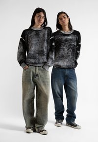Δύο μοντέλα φορούν υπερμεγέθη μαύρα και γκρίζα πουλόβερ με ριμπ μανσέτες και φαρδιές τζιν παντελόνες σε ανοιχτές και σκούρες αποχρώσεις, συνδυασμένα με μαύρα αθλητικά.