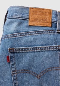 Plave traperice s tamno smeđom kožnom zakrpom s izraženim logotipom, kontrastnim narančastim šavovima i malim crvenim oznakom na stražnjem džepu.