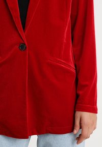 Blazer en velours rouge avec un seul bouton noir, deux poches avant, et manches longues, présentant une texture lisse et une coupe ajustée.