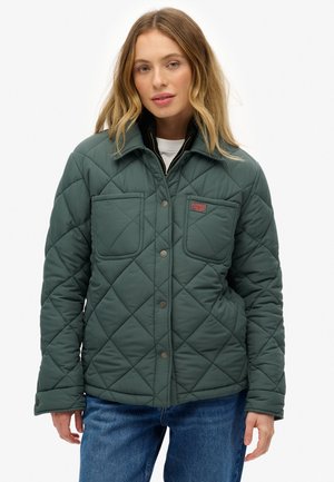 Veste matelassée verte avec fermeture à boutons, deux poches plaquées à l'avant, col et doublure intérieure contrastante. Portée sur une chemise blanche.