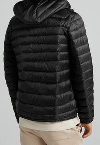 Muž stojící zády, oblečený v černé prošívané kapuci s horizontálními prošíváními, béžových kalhotách a světlebarevném tričku.