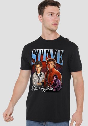 Young man wearing black T-shirt featuring multiple portraits of Steve Harrington with his name in large blue and white text.