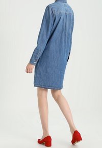 Světle modrá džínová košilová šaty s dlouhými rukávy, základním límcem, délkou do poloviny stehna, bočními rozparky a prošitým lemem. V kombinaci s červenými blokovými podpatky.