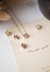 Gouden letters en charme kralen op een lichtbeige kaart. De kralen hebben reliëfletters en een hartvorm. Een fijne gouden ketting is zichtbaar.