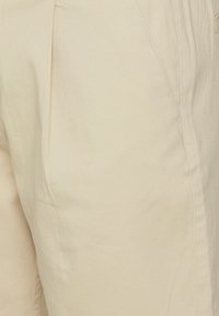 Beige chinos s uvolněným střihem, lehkou bavlněnou tkaninou, předními záhyby a bočními kapsami. Hladká textura a neutrální barva.