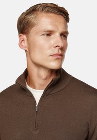 Brown zip-neck sweater made of textured fabric, featuring a standing collar and a silver zipper detail at the neck. Light-colored shirt underneath.