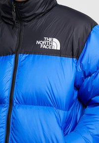 Veste matelassée bleue et noire avec un col montant, présentant un logo de marque, une fermeture éclair et une texture lisse et brillante.