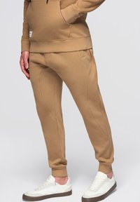 Personne portant un pantalon de jogging beige et un sweat à capuche assorti avec des baskets blanches, debout devant un fond clair uni.