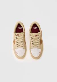 Beige og hvite Nike SB-sko med maroon fôr og små røde Nike swooshes på tåområdet, vist ovenfra på en hvit bakgrunn.