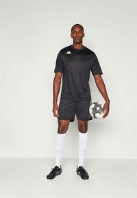 Μαύρη αθλητική φανέλα και σορτς με λευκό λογότυπο, λευκες κάλτσες μέχρι το γόνατο και μαύρες ποδοσφαιρικές μπότες, κρατώντας μια κανονική ποδοσφαιρική μπάλα.