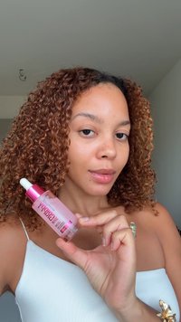 Flacon de sérum rose croustillant avec pipette, corps transparent et texte en gras. Cheveux bouclés avec une texture naturelle. Fond clair, distractions minimales.