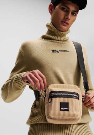 Beige Cordura-Crossbody-Tasche mit einem Reißverschluss, schwarzen Akzenten und einem Markenpatch. Kombiniert mit einem beigen gerippten Rollkragensweater.