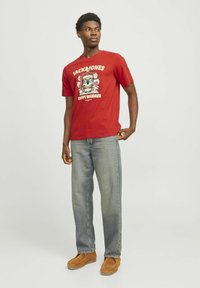 Camiseta de algodón roja con un estampado gráfico de un personaje y el texto "JACK&JONES HAPPY SHOKDAZE", combinada con jeans de mezclilla clara y zapatos marrón claro.