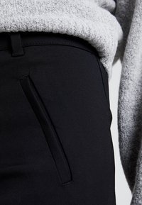 Svarta tygbyxor med en slät yta, utrustade med en sidoficka och en stilren design, kombinerade med en grå stickad tröja ovanför.