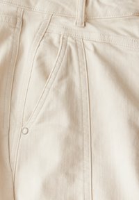 Tela de mezclilla beige claro con costuras visibles y un detalle de bolsillo delantero, que presenta un acento de remache metálico plateado. Textura suave.