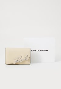 Beige leren portemonnee met zilveren "Karl" logo, voorzien van een flapsluiting. Vergezeld van een witte doos met zwarte tekst "KARL LAGERFELD."