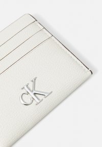 Tarjetero de cuero blanco con acabado texturizado, seis compartimentos para tarjetas y un acento del logo CK en plata en la parte frontal. Diseño minimalista con detalle de costura.